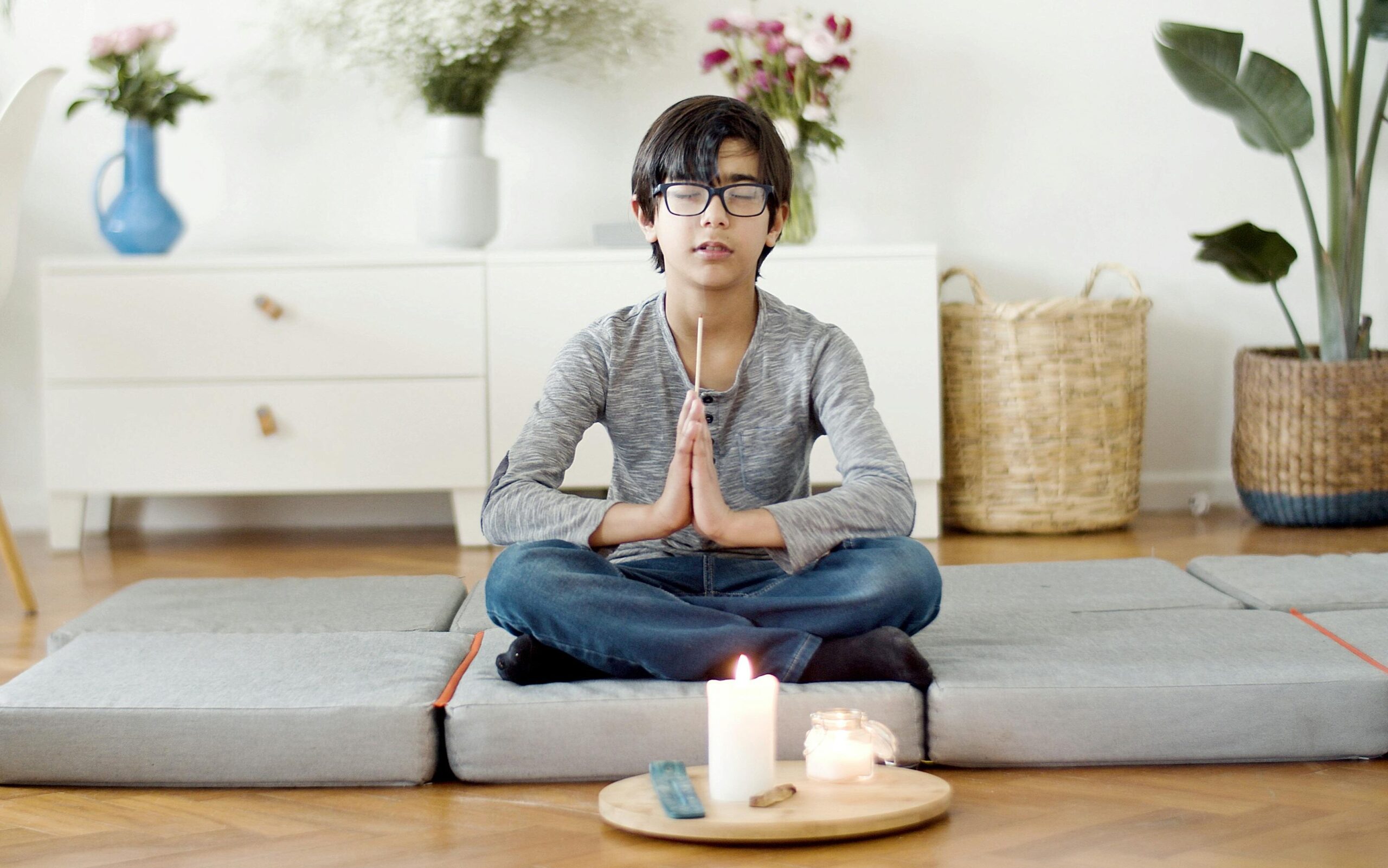 Young boy meditating at home with eyes closed, surrounded by candles and plants.