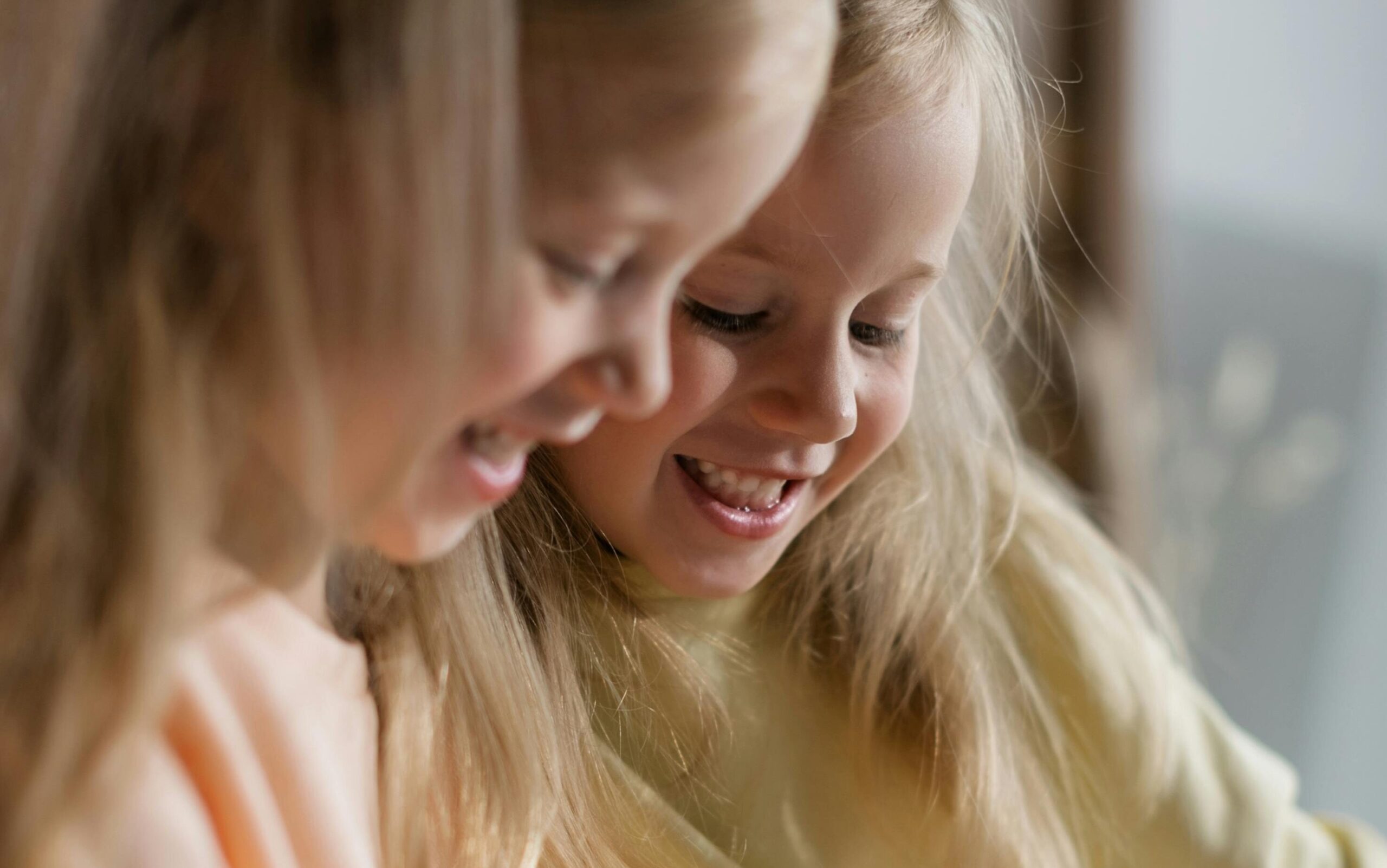Two smiling blonde sisters enjoying a happy moment indoors, showcasing sibling bonding.